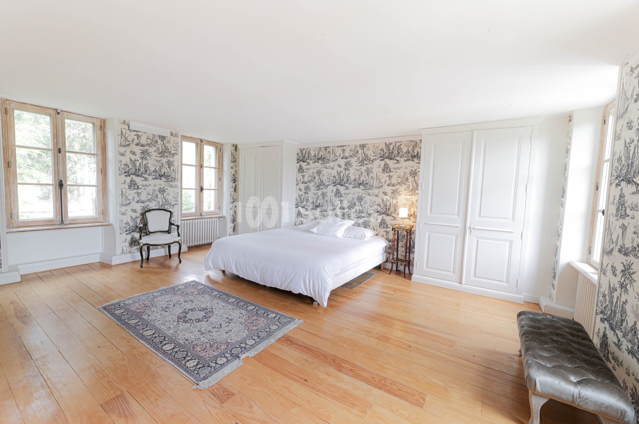 Chambre lumineuse avec lit double, parquet en bois, tapis, murs tapissés et grandes fenêtres donnant sur l'extérieur.