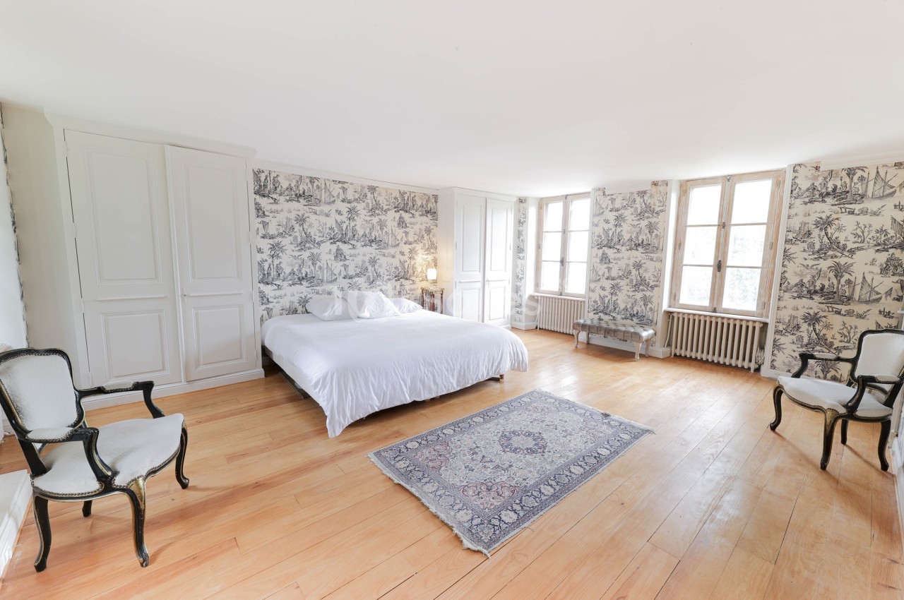 Chambre lumineuse avec lit double, parquet en bois, murs ornés de papier peint à motifs et fenêtres à double battant.