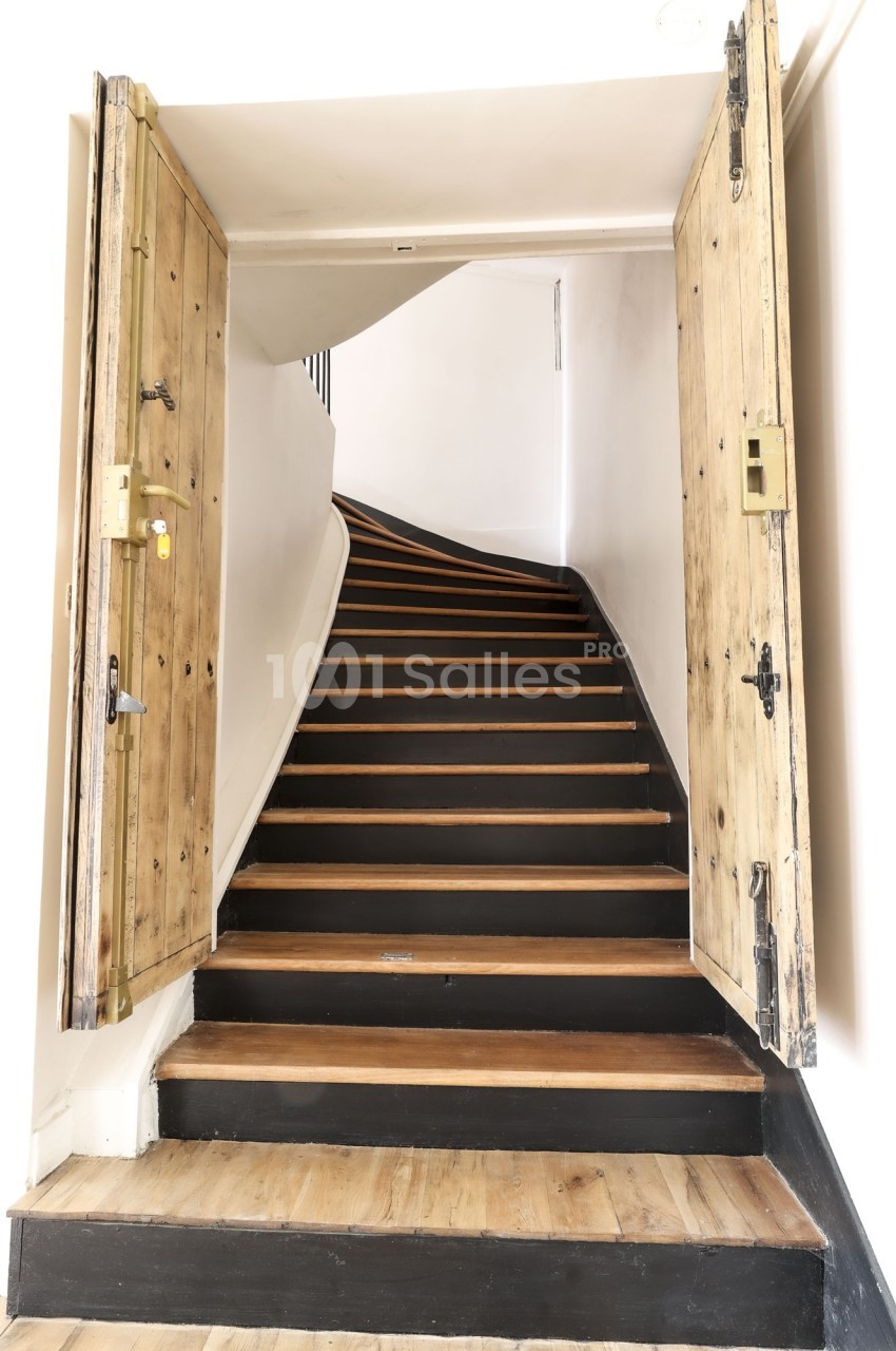Escalier en bois sombre avec des contremarches claires, encadré par des portes en bois brut ouvertes.