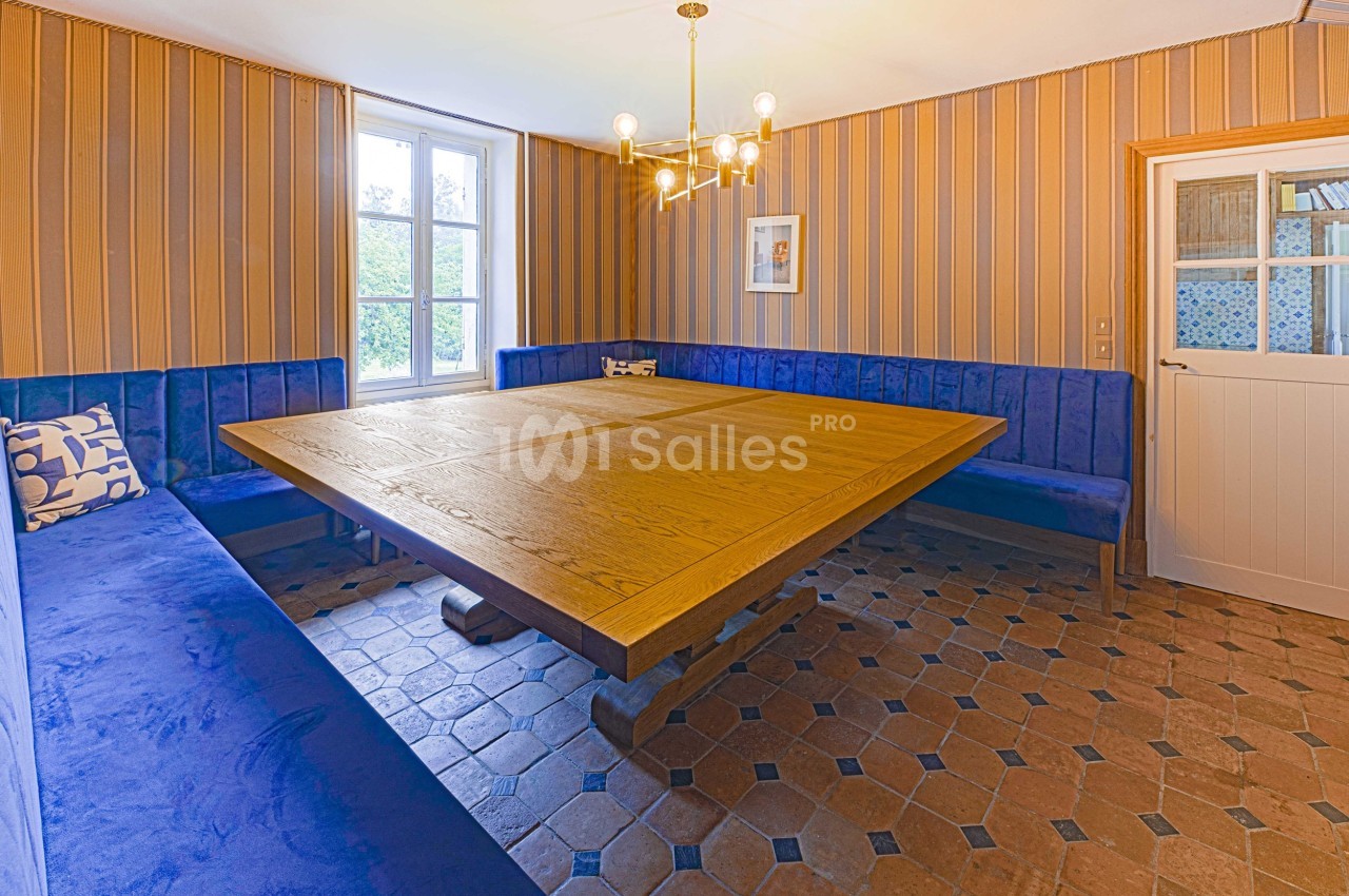 Salle à manger avec table en bois massif, banquettes bleues, carrelage ancien et lumière naturelle provenant des fenêtres.