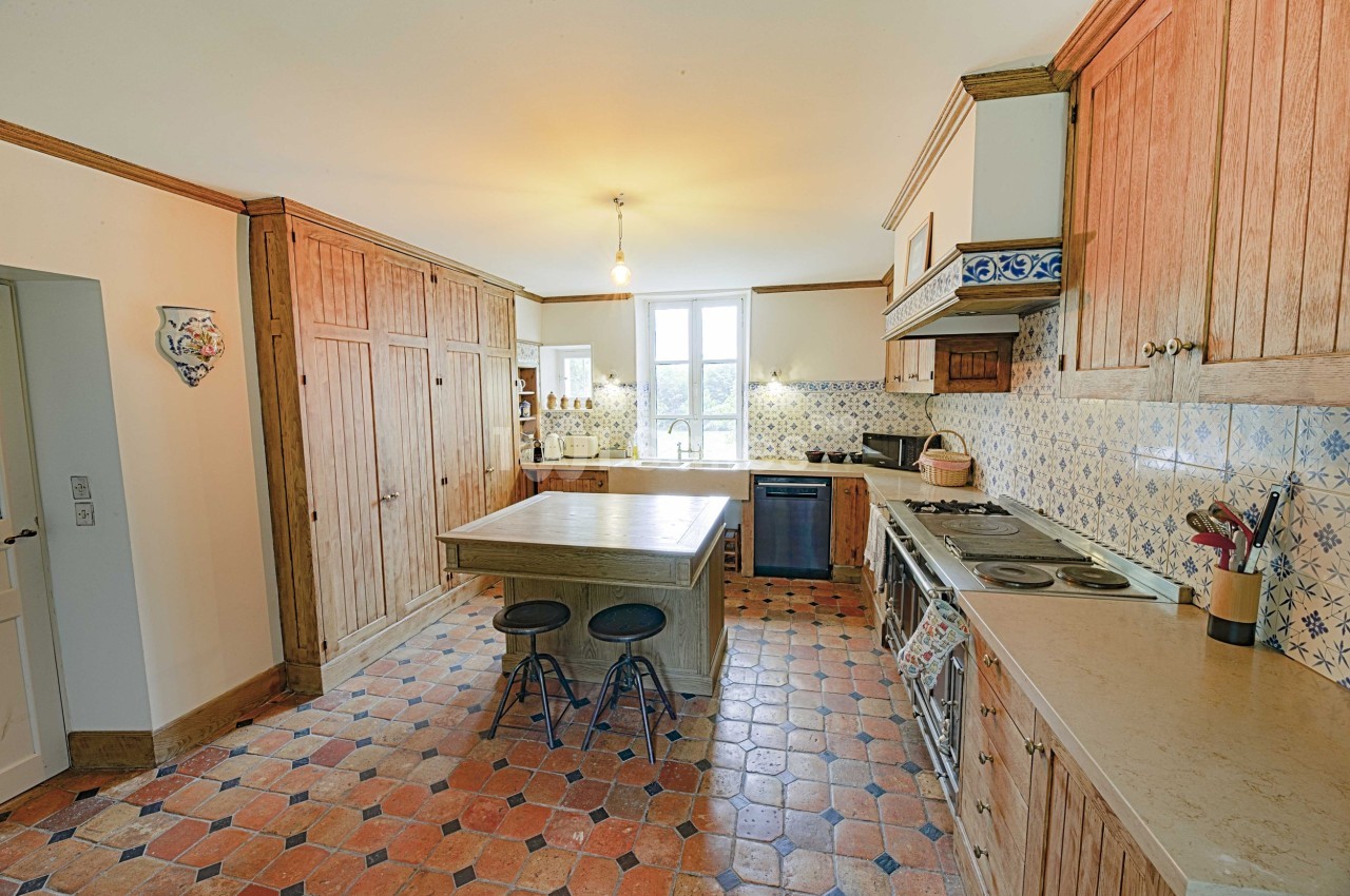Cuisine lumineuse avec meubles en bois, îlot central, carrelage en terre cuite et crédence à motifs.