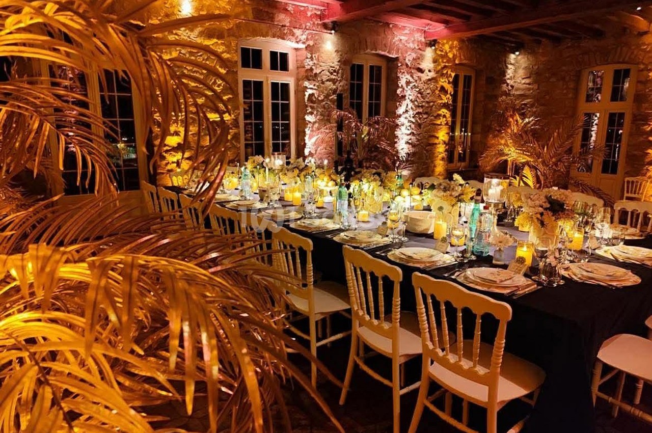 Salle de réception chaleureusement éclairée avec une grande table dressée, entourée de chaises blanches et de décorations…