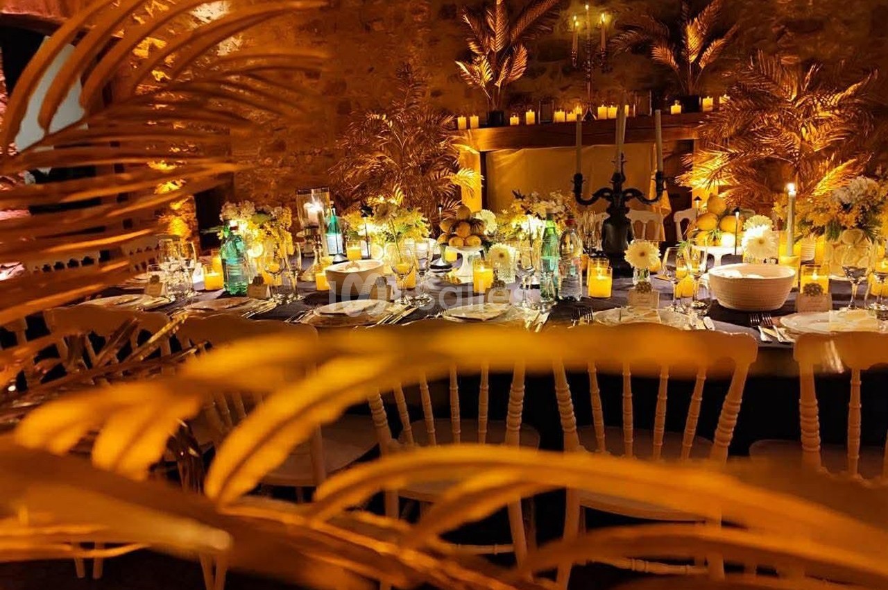 Table décorée pour un dîner élégant, entourée de bougies, fleurs et palmiers dorés dans une ambiance chaleureuse.
