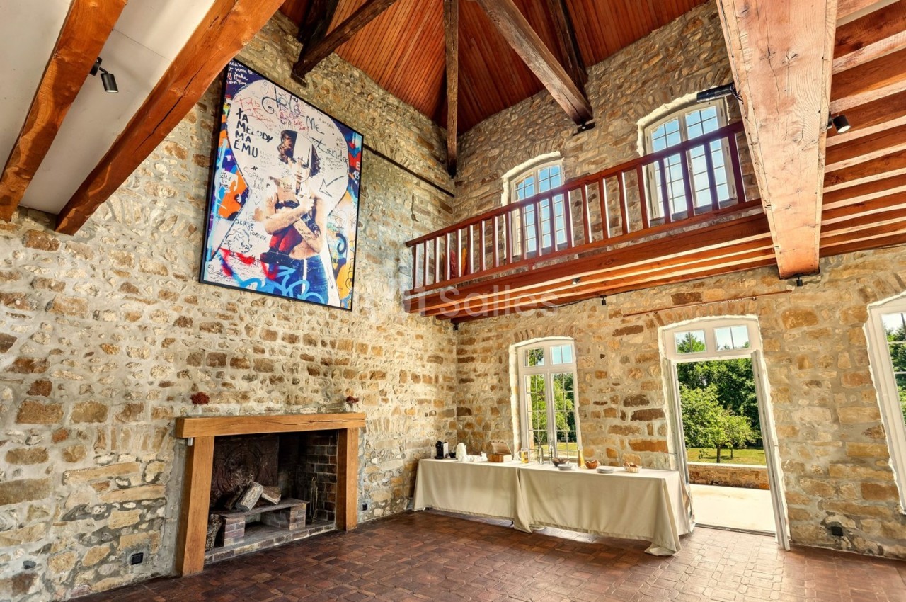 Salle en pierre avec cheminée, mezzanine en bois, tableau coloré au mur et table dressée près de grandes fenêtres.