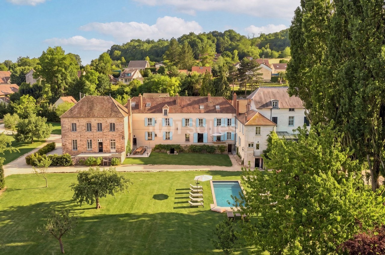 Vue aérienne d'une grande maison de campagne entourée d'un jardin verdoyant avec une piscine et des arbres.