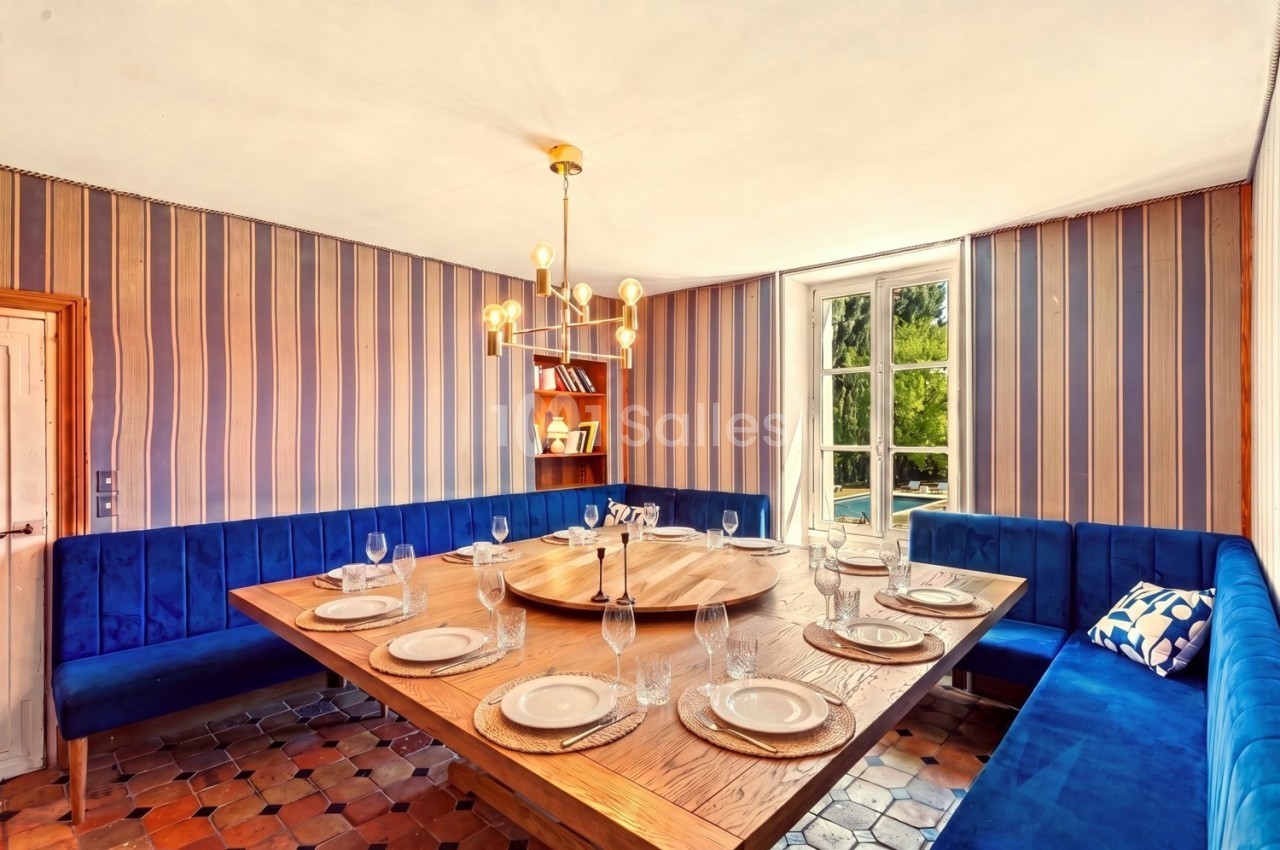 Salle à manger avec table en bois dressée, banquettes bleues, lustre moderne et fenêtre donnant sur un jardin.