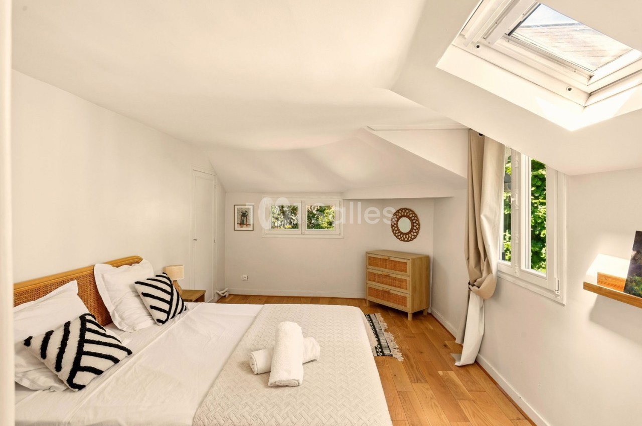 Chambre lumineuse avec lit double, parquet en bois, commode, fenêtres et puits de lumière donnant sur un extérieur verdoyant.