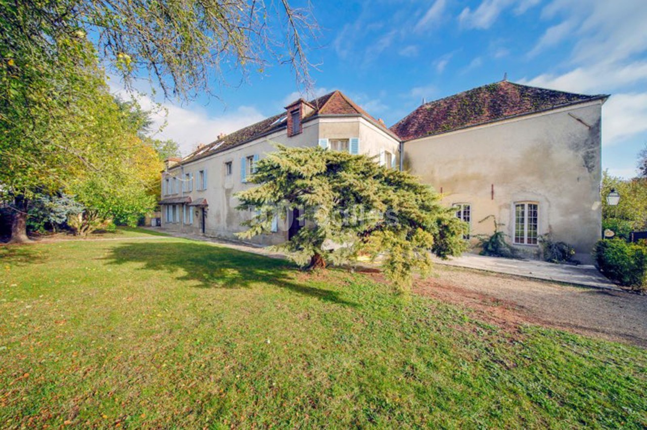 Grande maison ancienne avec toiture en tuiles, entourée d'un jardin arboré sous un ciel partiellement nuageux.