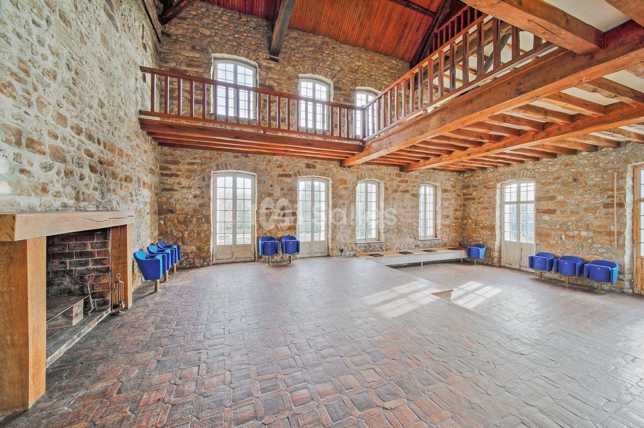 Salle spacieuse avec murs en pierre, plafond en bois, mezzanine, cheminée et grandes fenêtres laissant entrer la lumière.