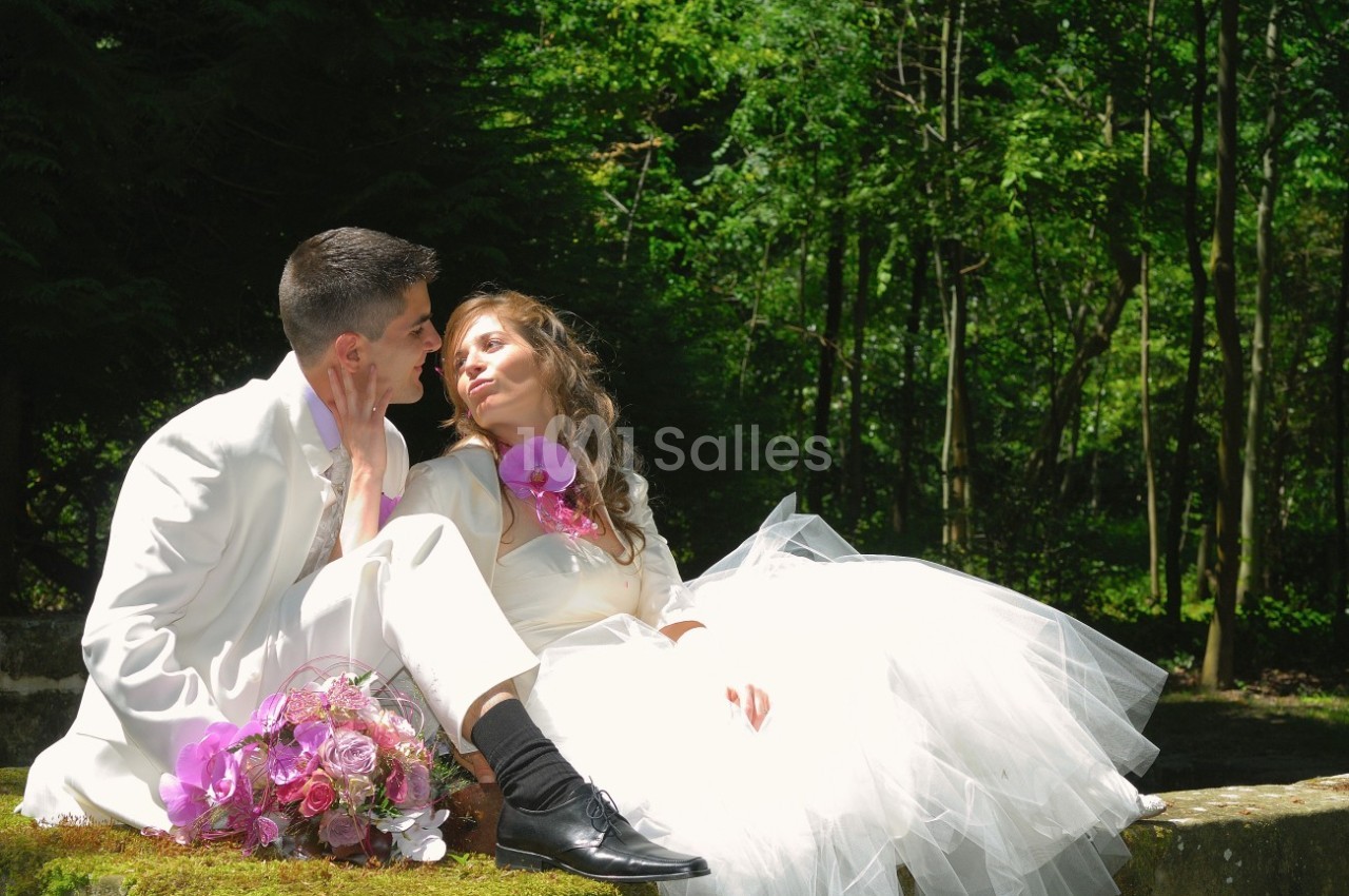 Un couple en tenue de mariage assis sur un muret, entouré de verdure, avec un bouquet de fleurs posé à côté.