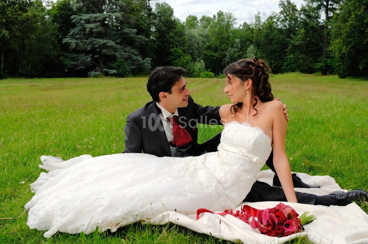 Un couple en tenue de mariage est assis dans un parc verdoyant, entouré de nature, avec un bouquet de fleurs rouges.
