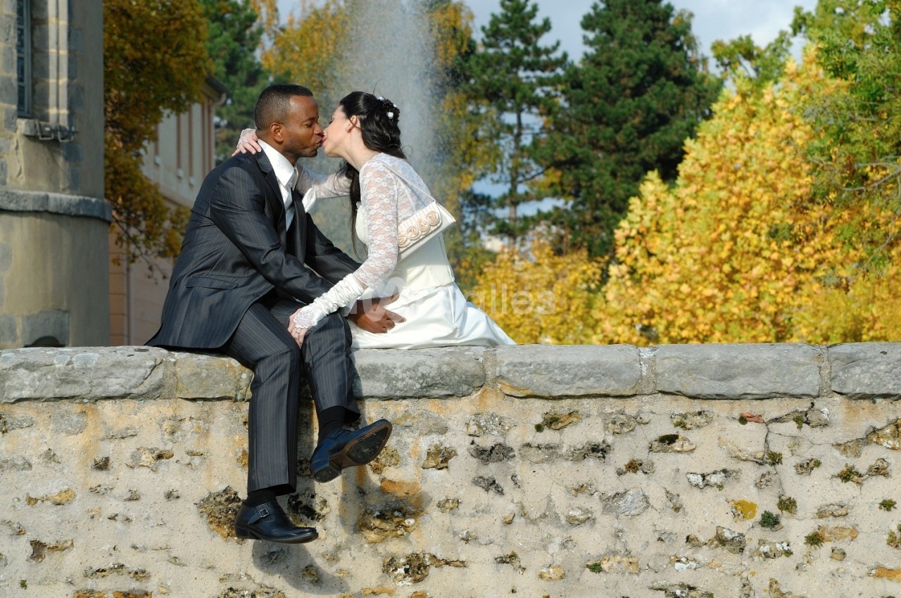 Un couple de mariés assis sur un muret en pierre, s'embrassant devant une fontaine et des arbres en automne.