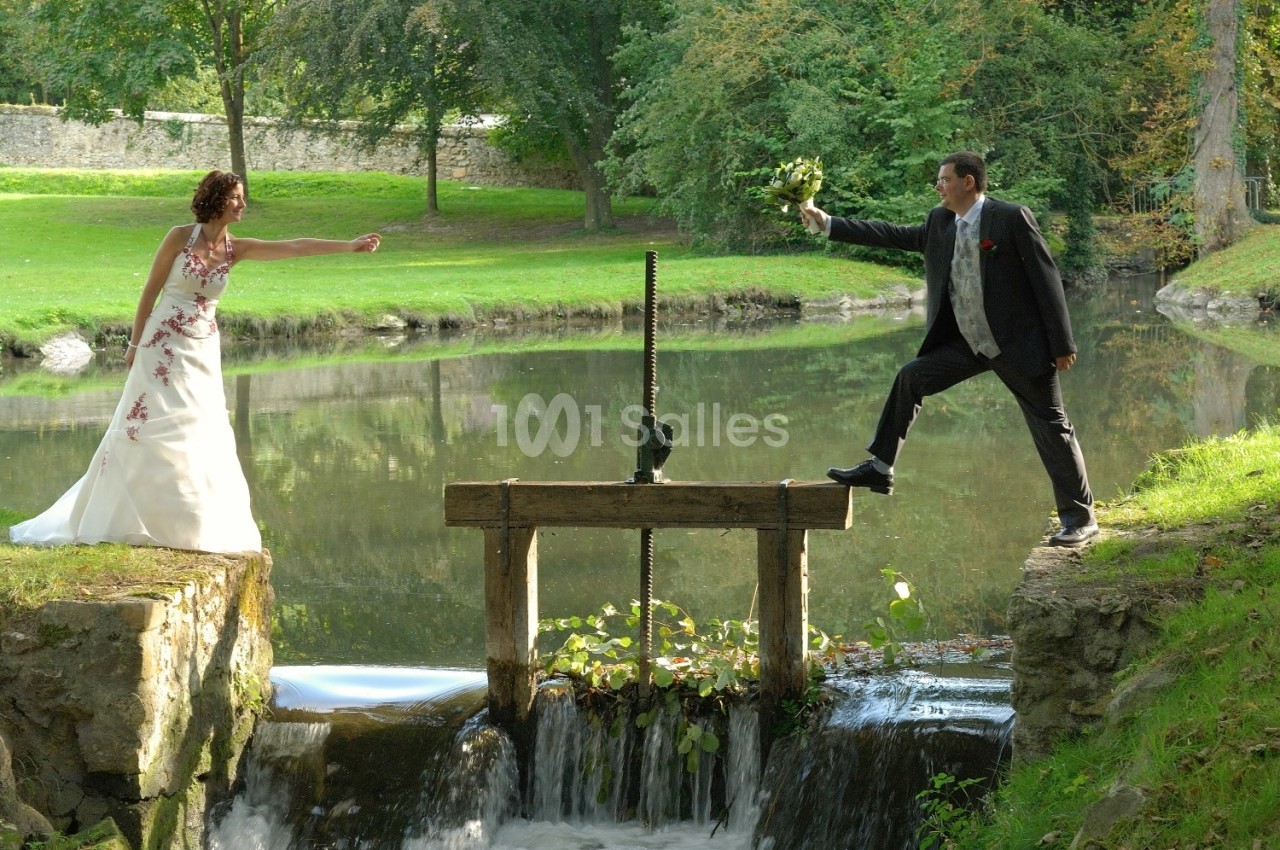 Un couple en tenue de mariage se tient de part et d'autre d'une petite écluse dans un cadre naturel verdoyant.