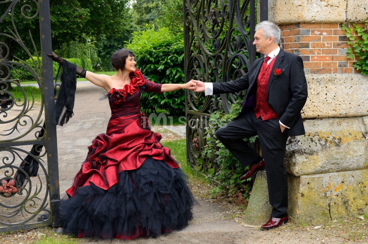 Un couple élégant en tenue de soirée pose devant une porte en fer forgé dans un jardin verdoyant.