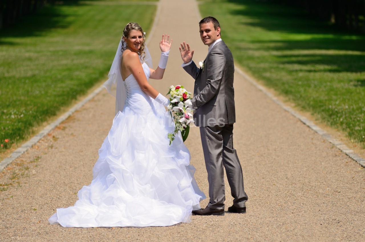 Un couple de mariés souriant, en tenue de cérémonie, se tient sur une allée bordée de pelouse, saluant vers l'objectif.