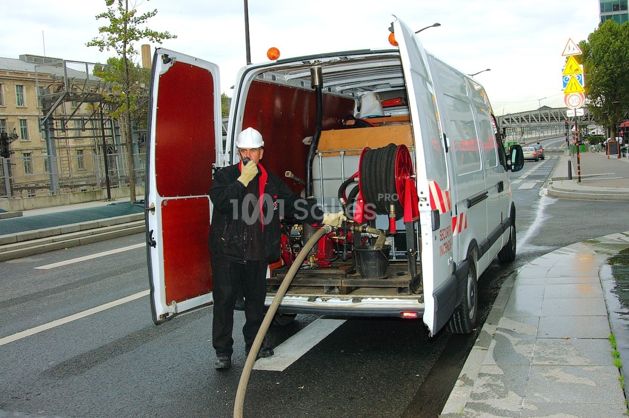 Un technicien en tenue de sécurité utilise un tuyau relié à un fourgon équipé de matériel technique sur une route.