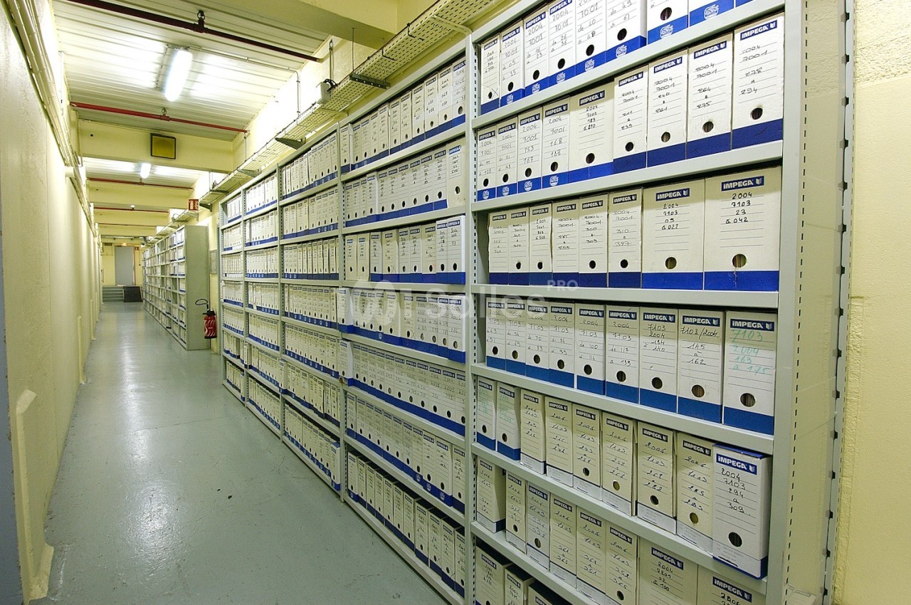 Rayonnages métalliques alignés contenant des classeurs d'archives bleus et blancs dans un couloir éclairé.