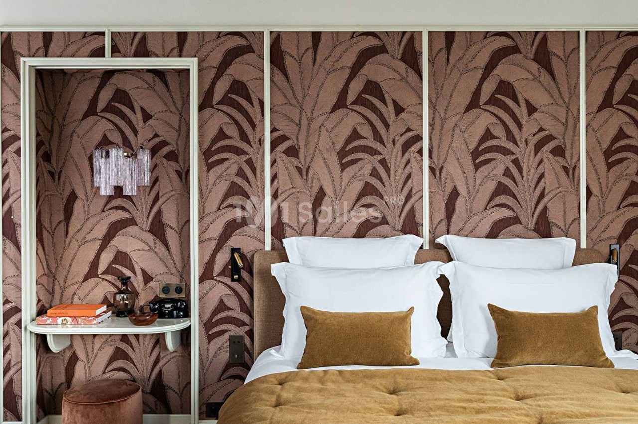 Chambre avec lit double, coussins marron, murs décorés de motifs végétaux et petite étagère avec objets.