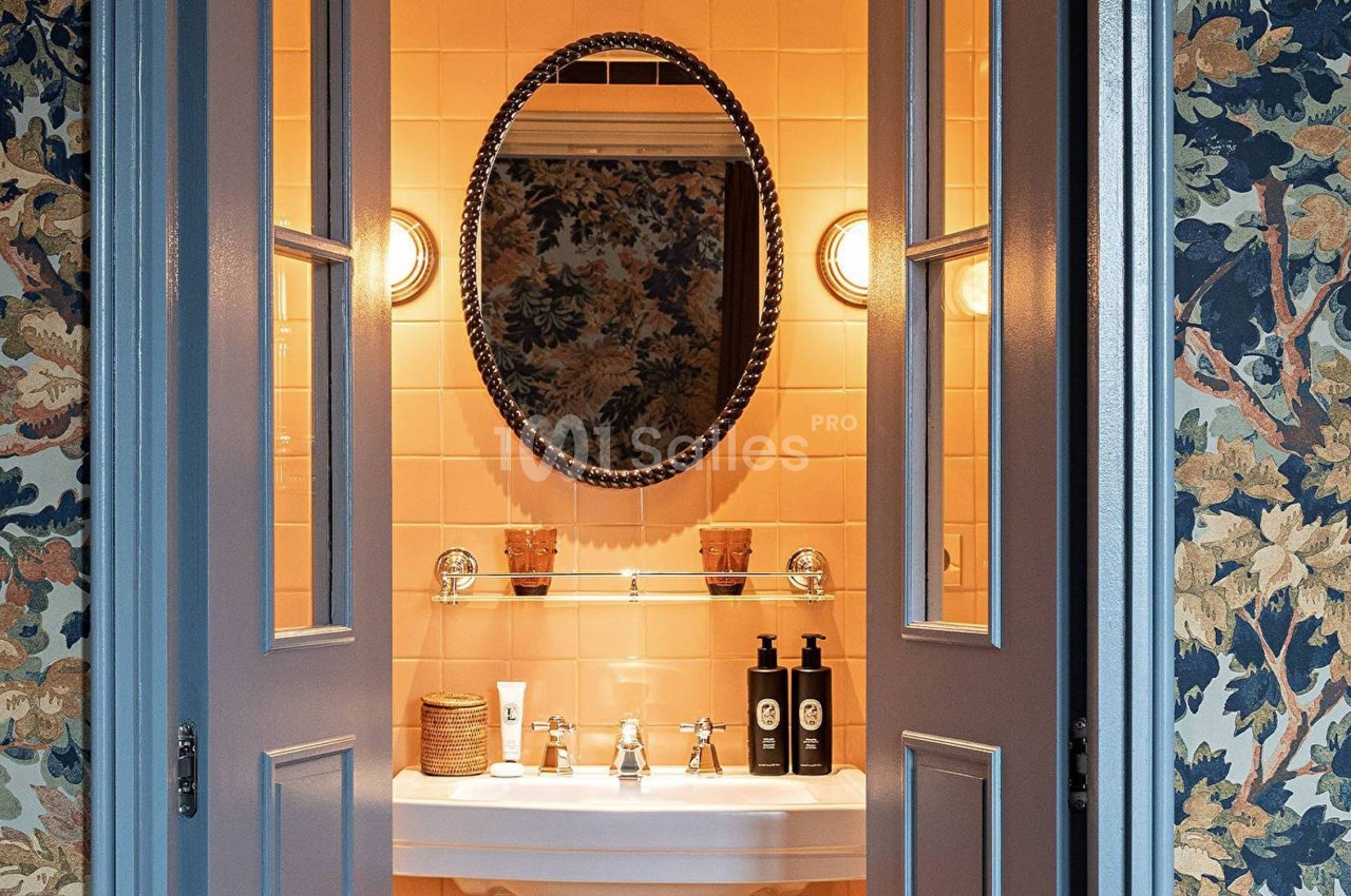 Lavabo avec miroir ovale, éclairage mural et décor floral sur les murs et carrelage beige en arrière-plan.