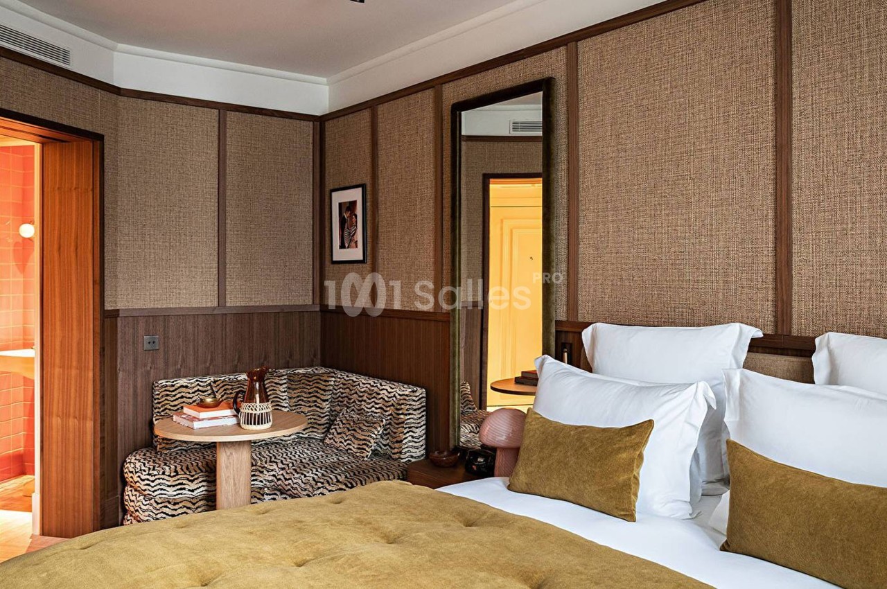 Chambre d'hôtel avec lit double, coussins jaunes, canapé imprimé, table basse et vue partielle sur une salle de bain.