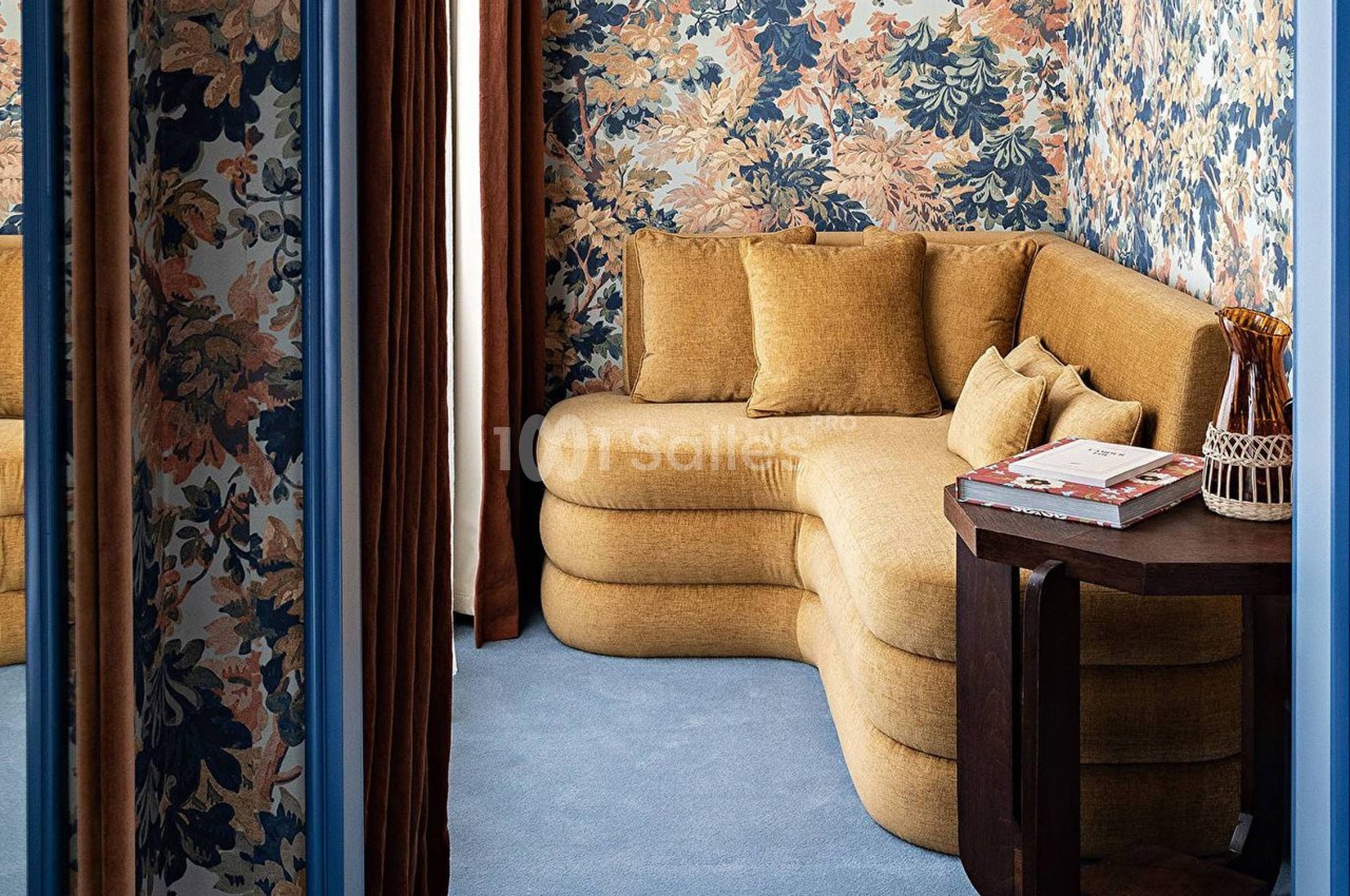 Canapé d'angle jaune moutarde dans une pièce aux murs tapissés de motifs floraux, avec une table en bois à côté.