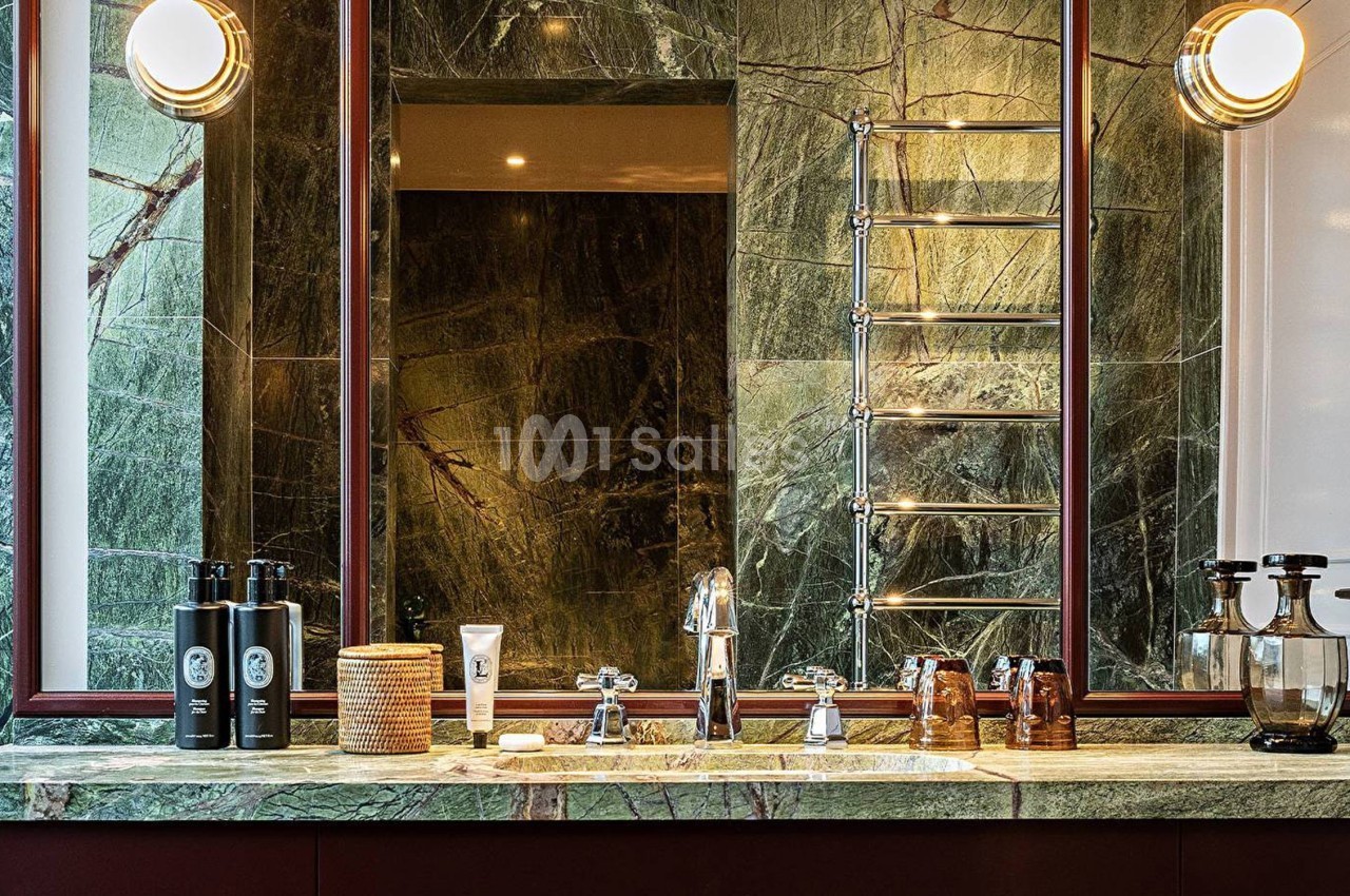 Salle de bain luxueuse avec un plan de travail en marbre vert, miroir encadré, produits de toilette et éclairage mural.