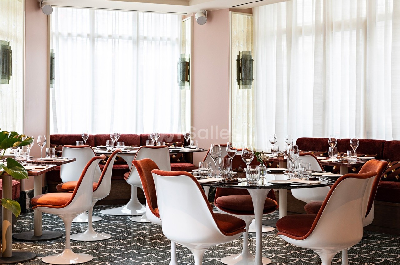 Salle de restaurant lumineuse avec tables dressées, chaises blanches et banquettes rouges, décorée de plantes et de rideaux.
