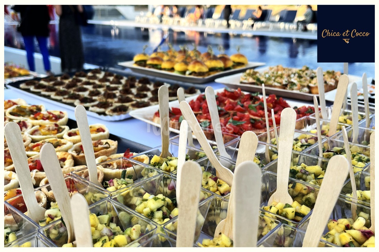 Buffet coloré avec verrines de salade, mini-burgers et assortiments de bouchées devant une piscine.