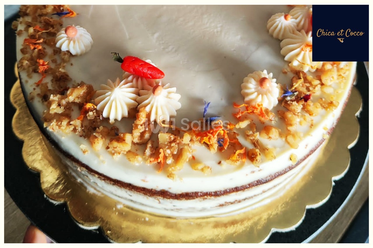 Gâteau décoré de crème, noix concassées, fleurs comestibles et un petit piment rouge sur un plateau doré.