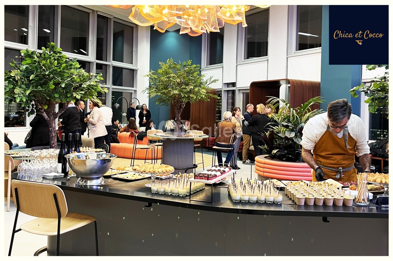Buffet dressé dans un espace moderne avec des invités debout en arrière-plan, discutant près de plantes décoratives.