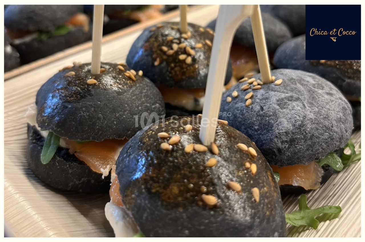 Mini-burgers au pain noir garnis de saumon fumé, roquette et fromage, présentés avec des piques en bois.