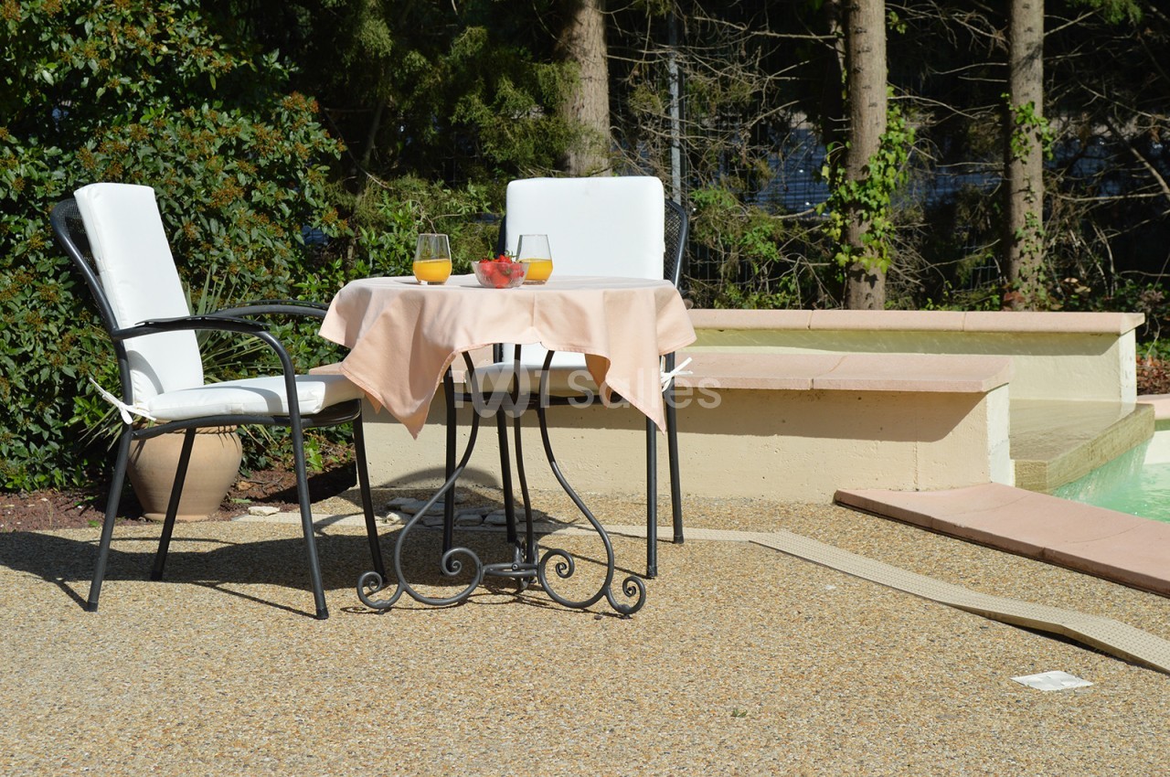 Table ronde avec nappe beige, deux chaises blanches, verres de jus d'orange et fraises, disposée près d'une piscine.
