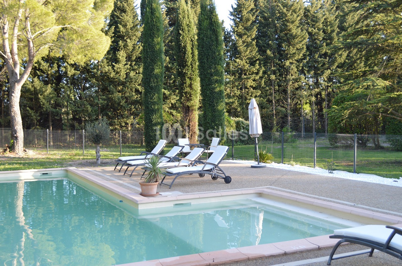 Piscine extérieure entourée de chaises longues, parasol et arbres, dans un cadre naturel verdoyant.