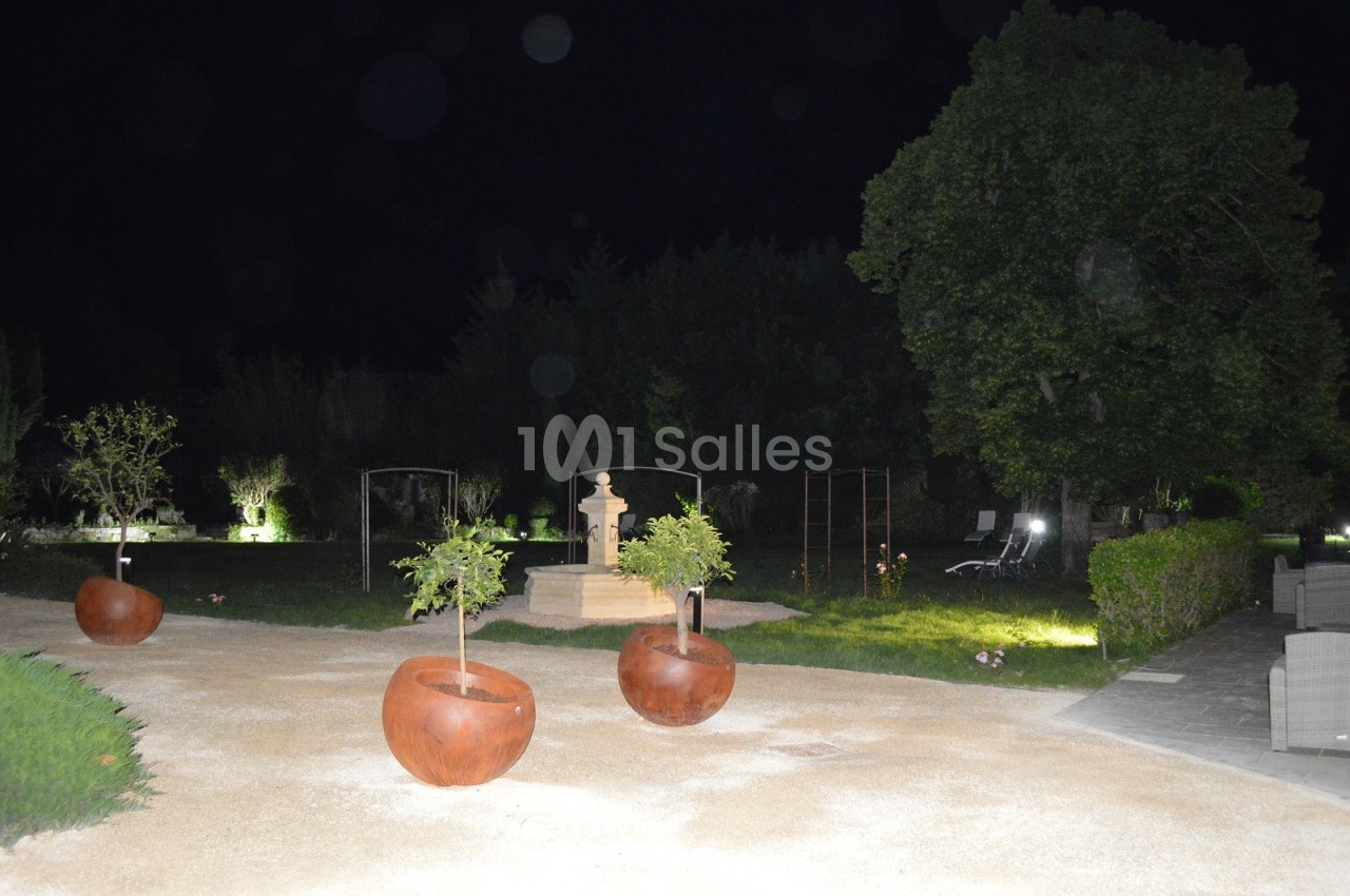 Allée de jardin éclairée la nuit, avec des pots en métal contenant de petits arbres et une fontaine en arrière-plan.