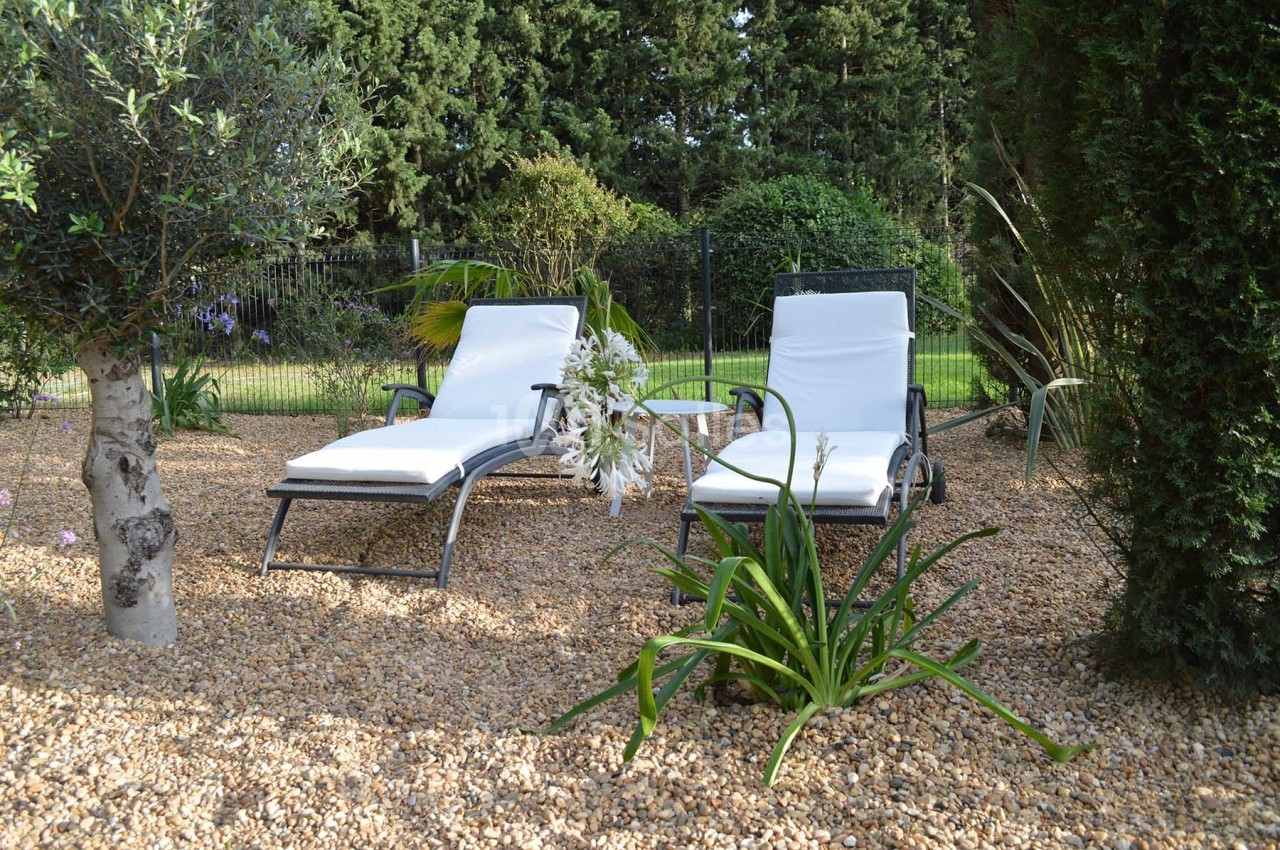 Deux chaises longues blanches sur un sol en gravier, entourées de plantes et d'arbres dans un jardin.