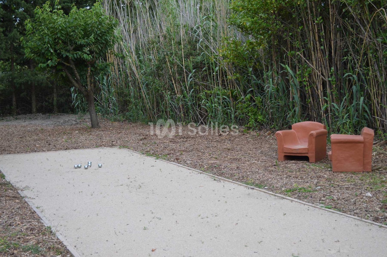 Terrain de pétanque en plein air entouré de végétation, avec deux fauteuils en terre cuite à proximité.