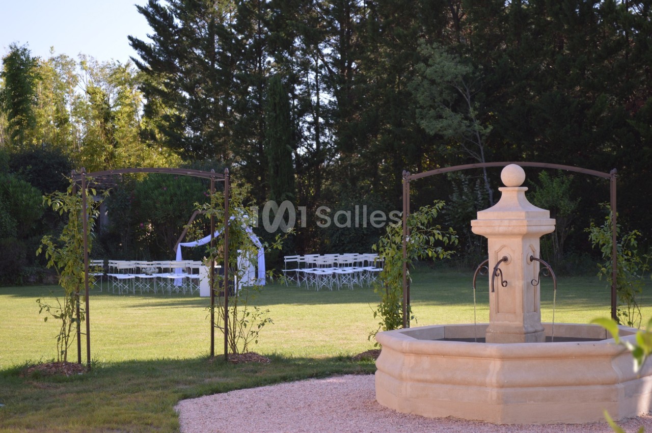 Fontaine en pierre au premier plan dans un jardin verdoyant, avec des chaises blanches disposées sous une arche au fond.