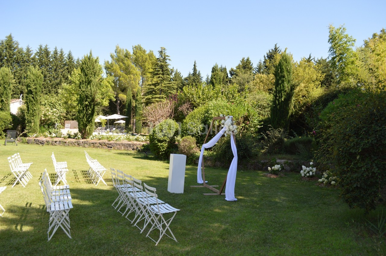 Chaises blanches disposées en extérieur devant une arche décorée, entourées de verdure et d'arbres.