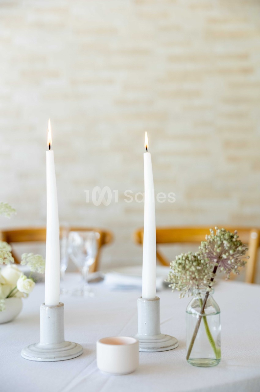 Deux bougies blanches allumées sur des chandeliers, avec un vase de fleurs et une table dressée en arrière-plan.