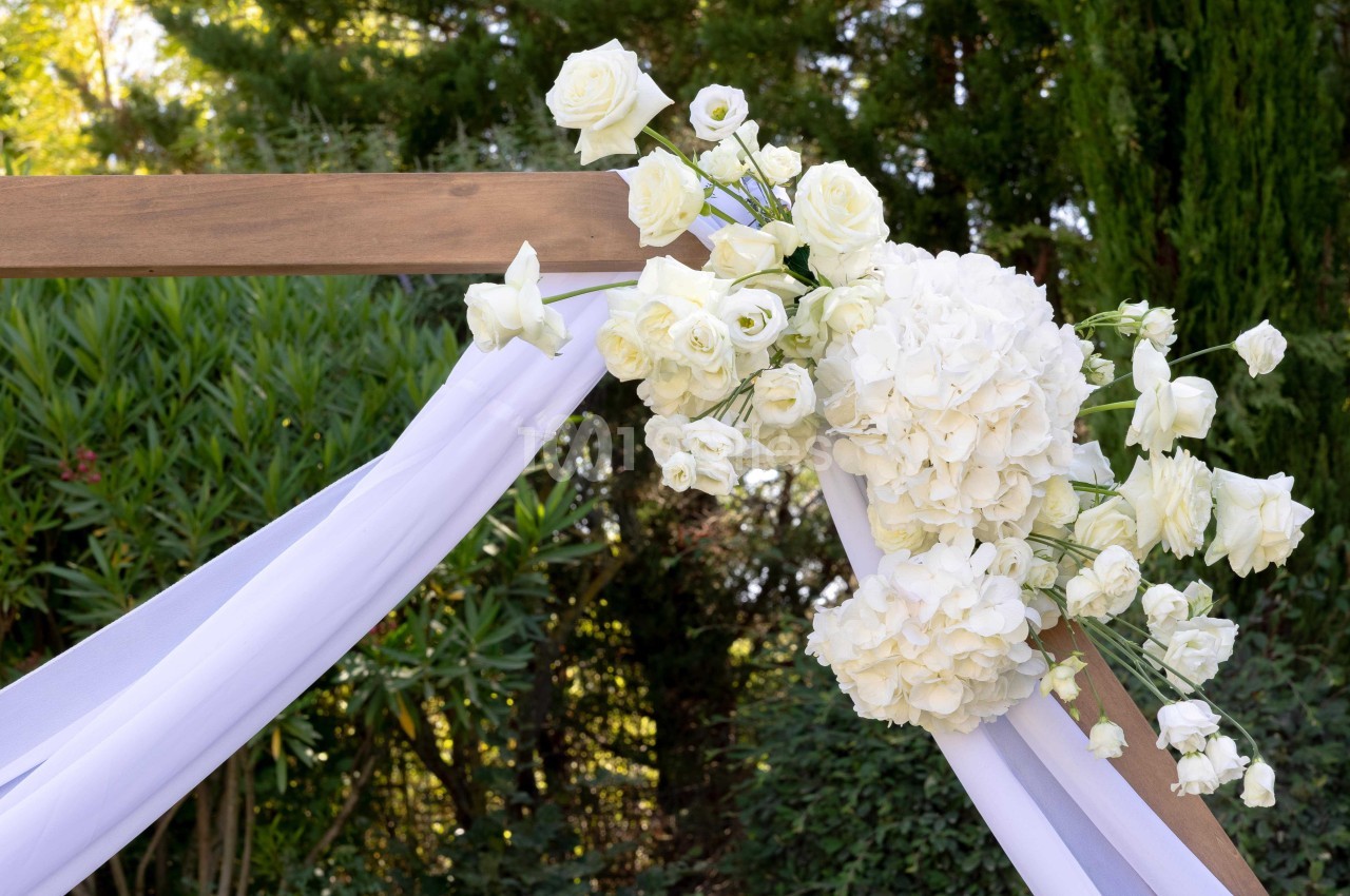 Arrangement floral blanc avec des roses et des hortensias, fixé sur une arche en bois décorée de voiles blancs, en extérieur.