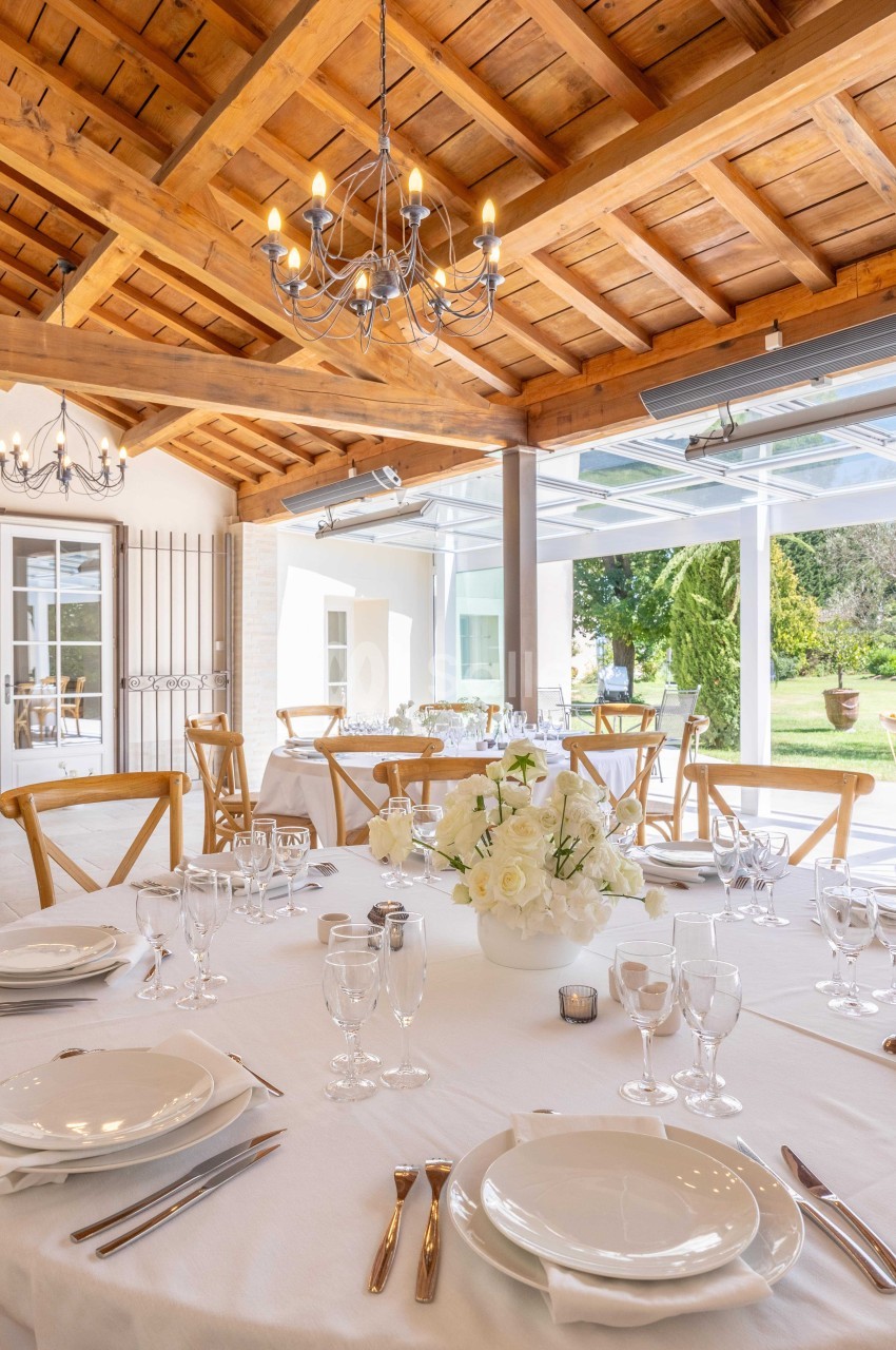 Salle lumineuse avec tables dressées, nappes blanches, vaisselle élégante, bouquet de fleurs et plafond en bois.