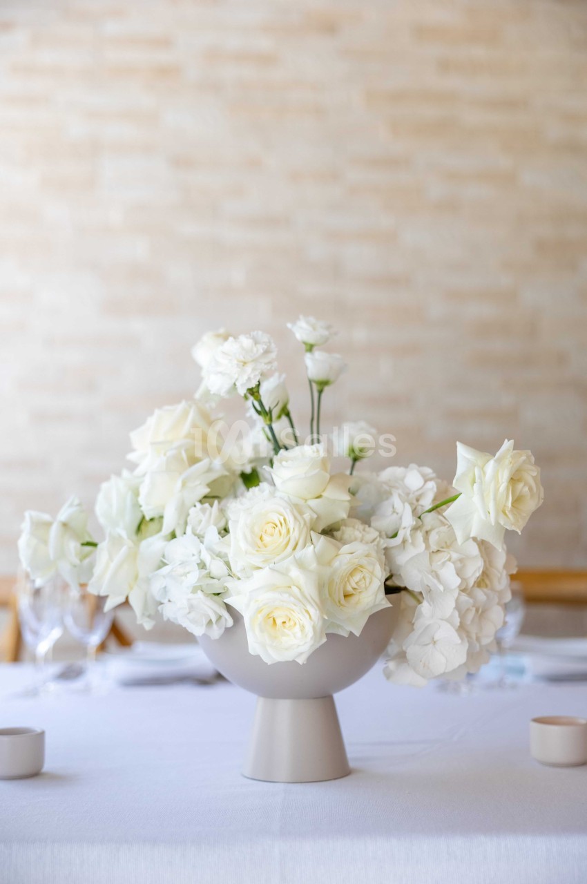 Arrangement de fleurs blanches, incluant des roses et des hortensias, dans un vase beige sur une table blanche.