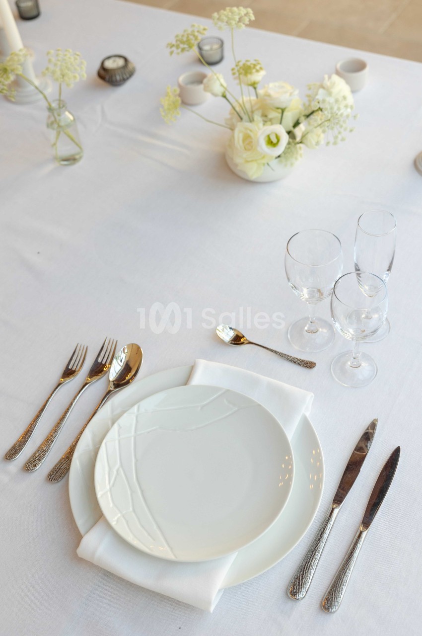Table dressée avec assiette blanche, couverts en métal, serviette pliée, verres à pied et centre de table floral.
