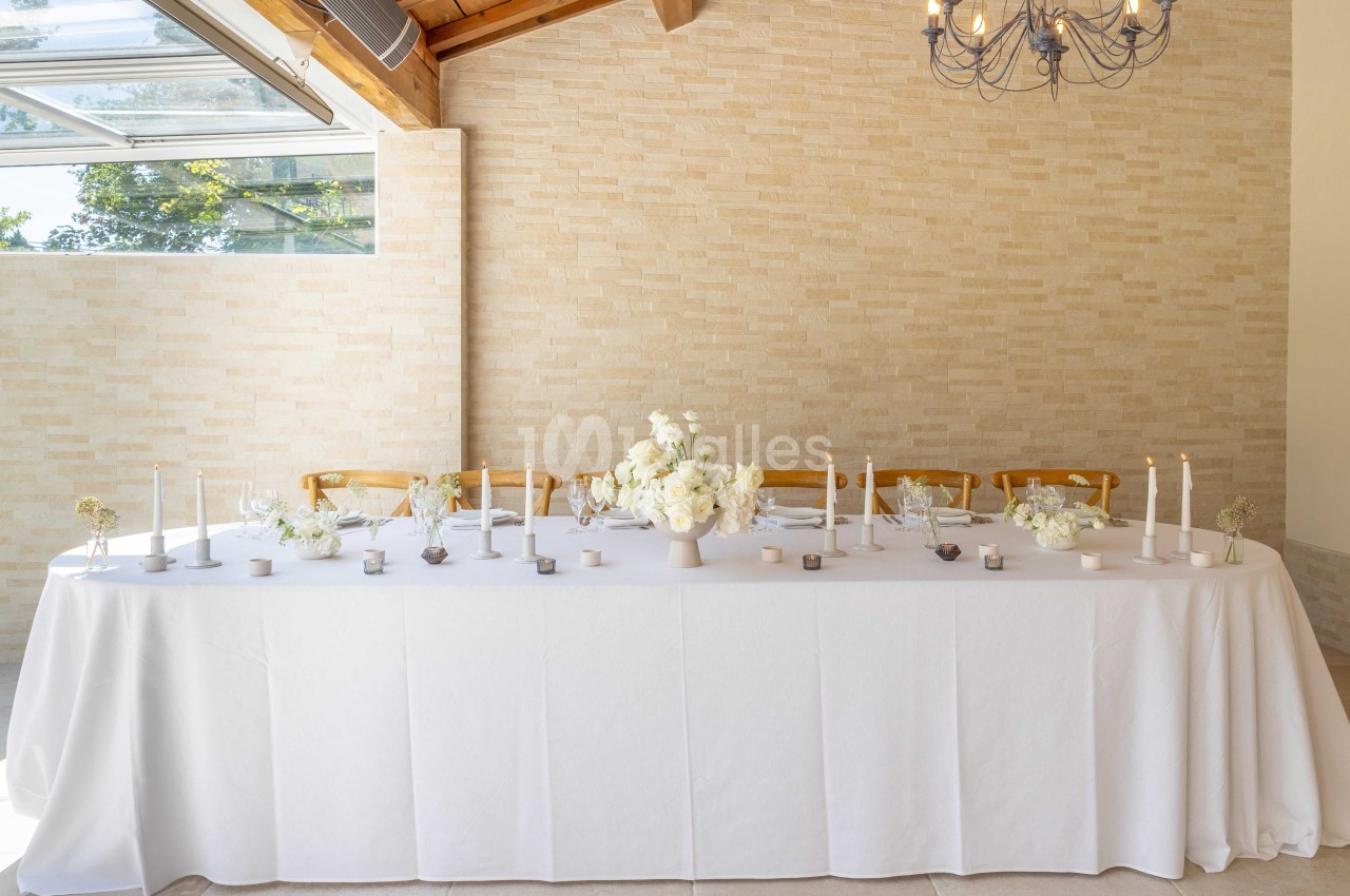 Table décorée avec une nappe blanche, des bougies, des fleurs blanches et des chaises en bois dans une salle lumineuse.