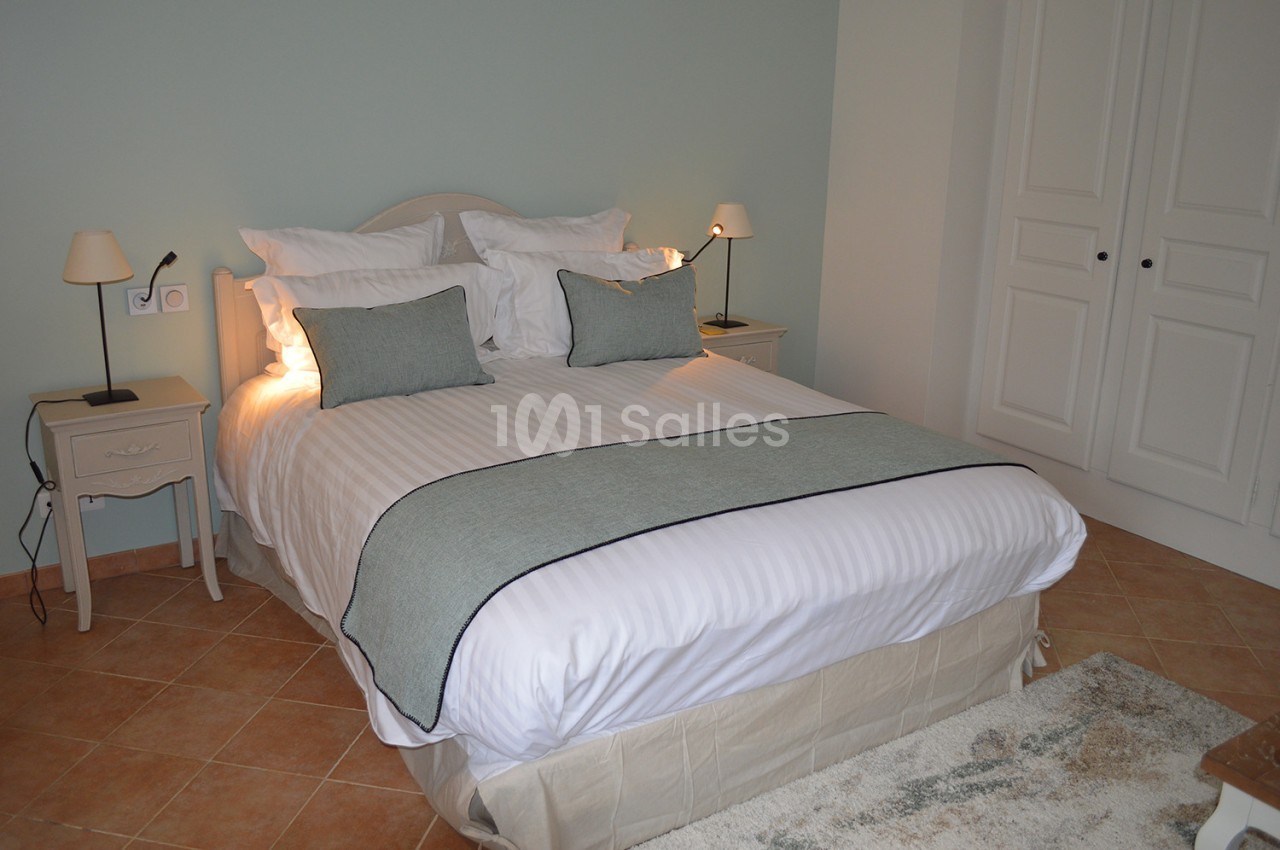 Lit double avec draps blancs et couvre-lit gris, encadré par deux tables de chevet avec lampes, dans une chambre lumineuse.