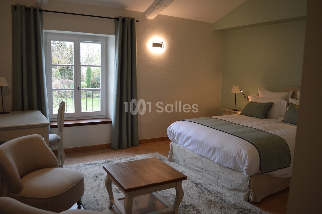Chambre lumineuse avec lit double, bureau, fauteuils et vue sur un jardin à travers une fenêtre.