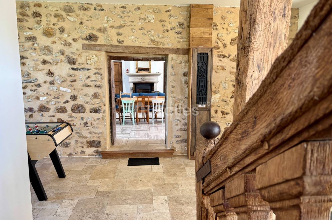 Vue intérieure d'une maison en pierre avec un baby-foot, un escalier en bois et une ouverture vers une salle à manger.