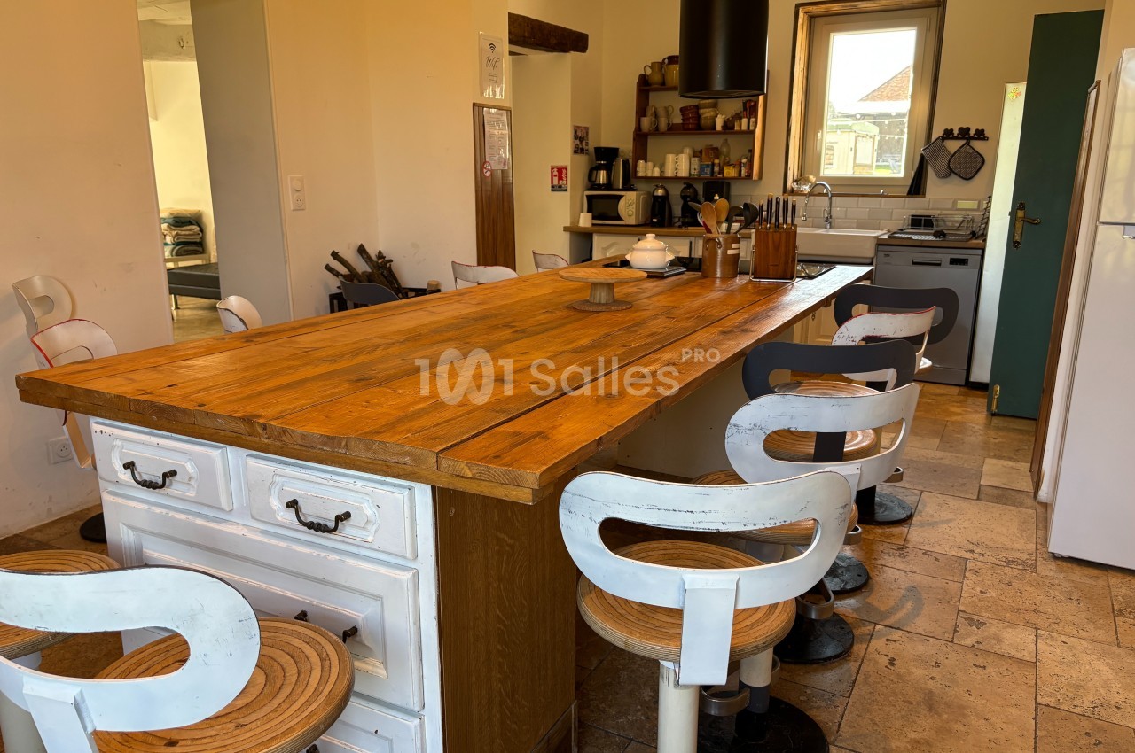 Cuisine lumineuse avec une grande table en bois, des chaises blanches, des étagères et des appareils électroménagers.