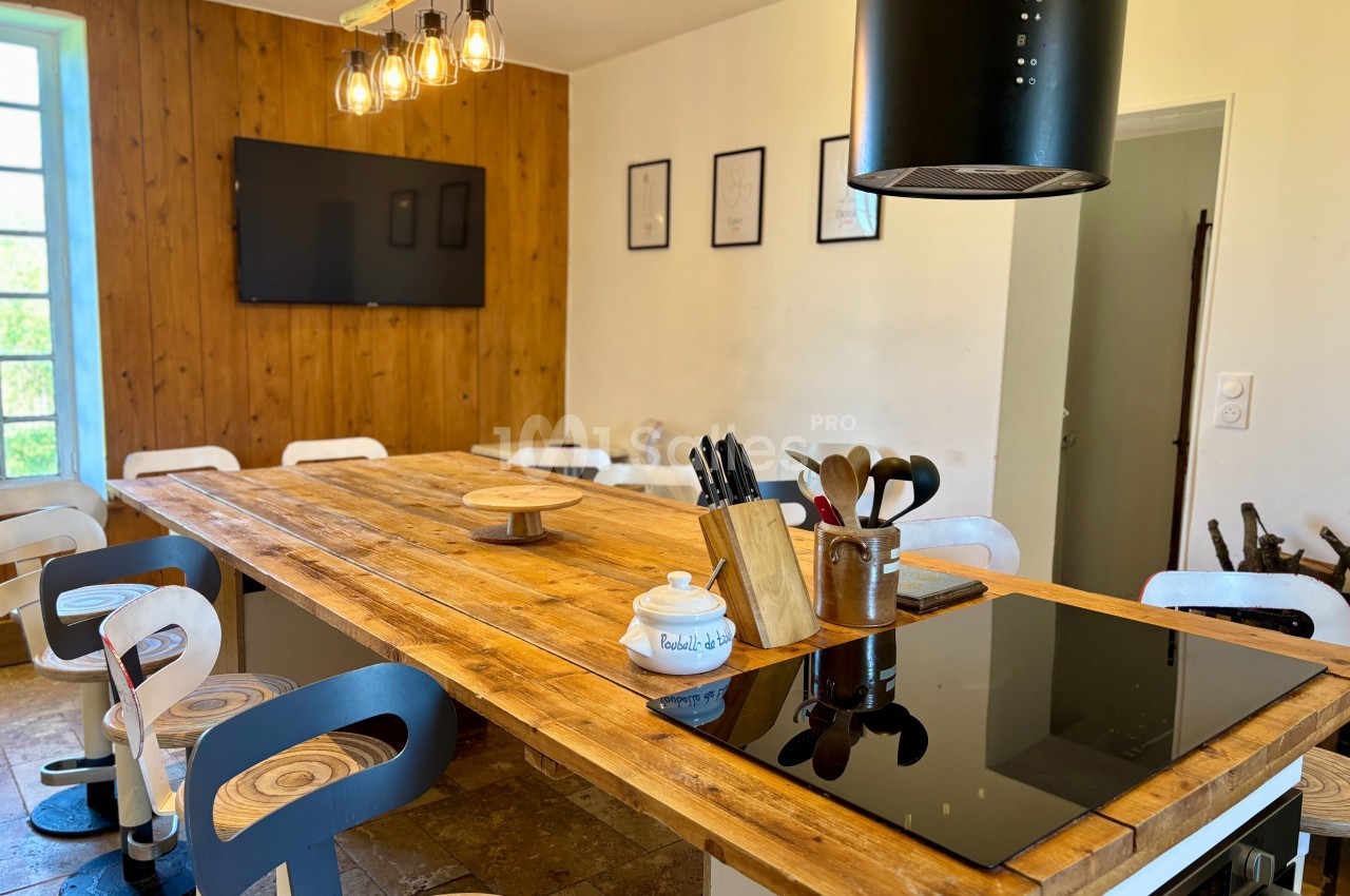 Grande table en bois entourée de tabourets dans une cuisine lumineuse avec plaques de cuisson et décorations murales.