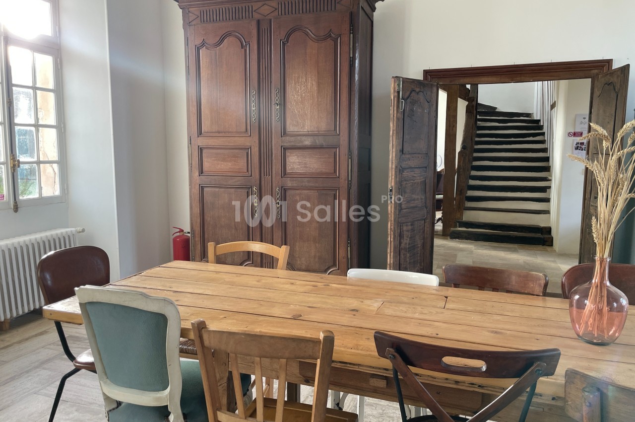 Salle lumineuse avec une grande table en bois entourée de chaises dépareillées, une armoire ancienne et un escalier en…